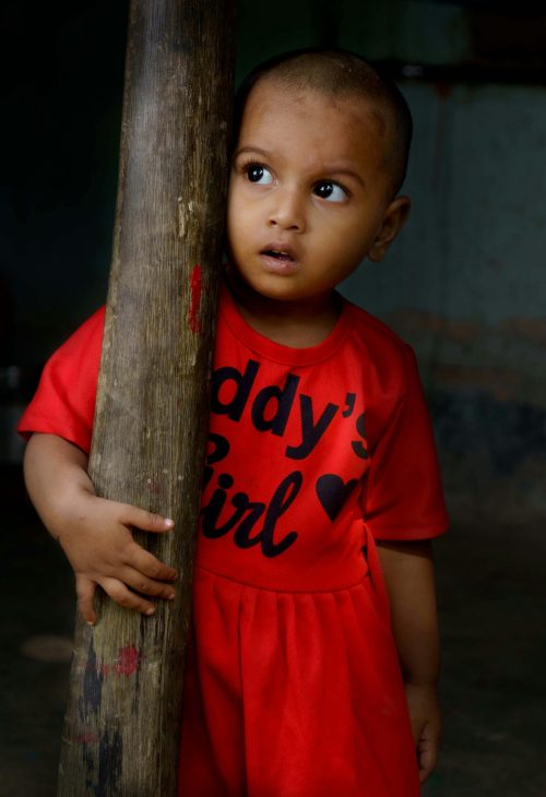Adorable child in red dress indoors in Pingla, India, holding a wooden post.